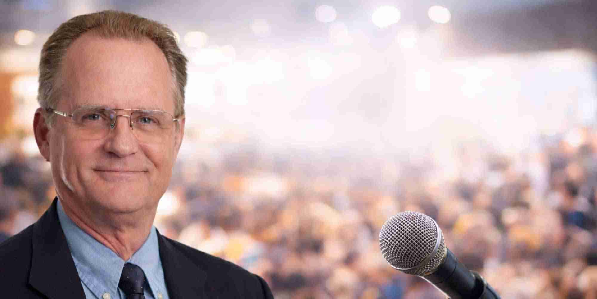 John Tooley speaking at a conference with microphone and audience in background